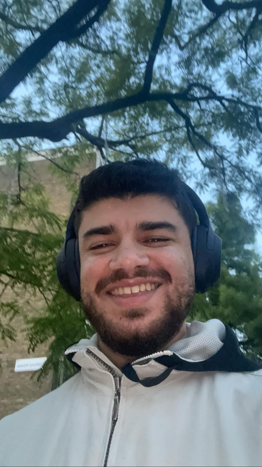 Man smiling outdoors wearing headphones and a white jacket, with trees in the background.