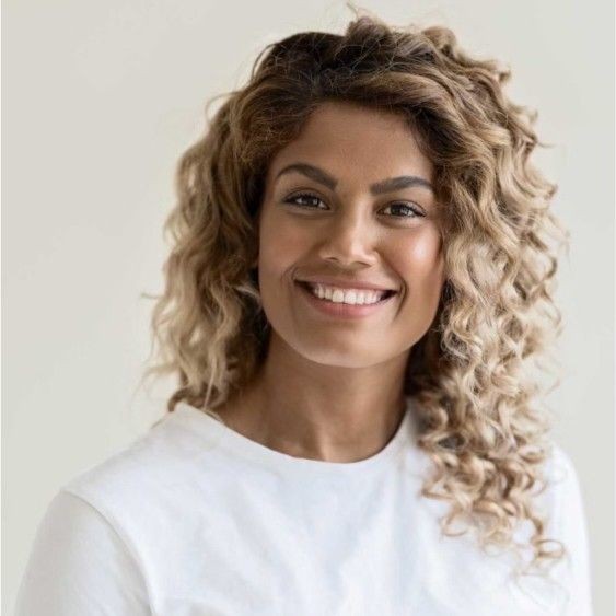 Smiling person with curly hair wearing a white shirt, set against a plain background.