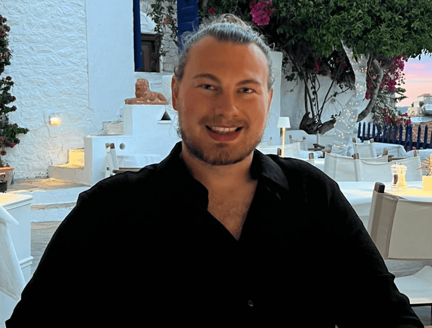 Smiling person in a black shirt sitting in an outdoor restaurant with white decor and greenery.