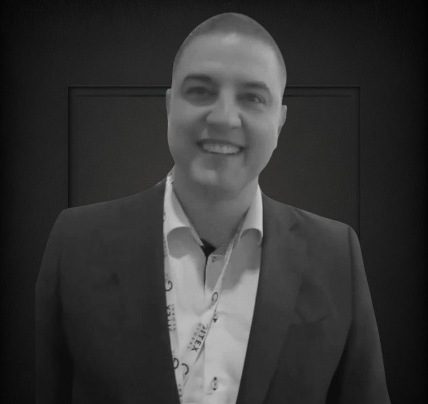 Smiling person in a suit with a lanyard, against a dark background.