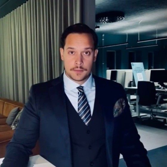 Man in a suit standing in an office with computers and a curtain backdrop.