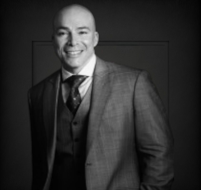 Man wearing a grey suit with a patterned tie and vest, standing against a dark background.