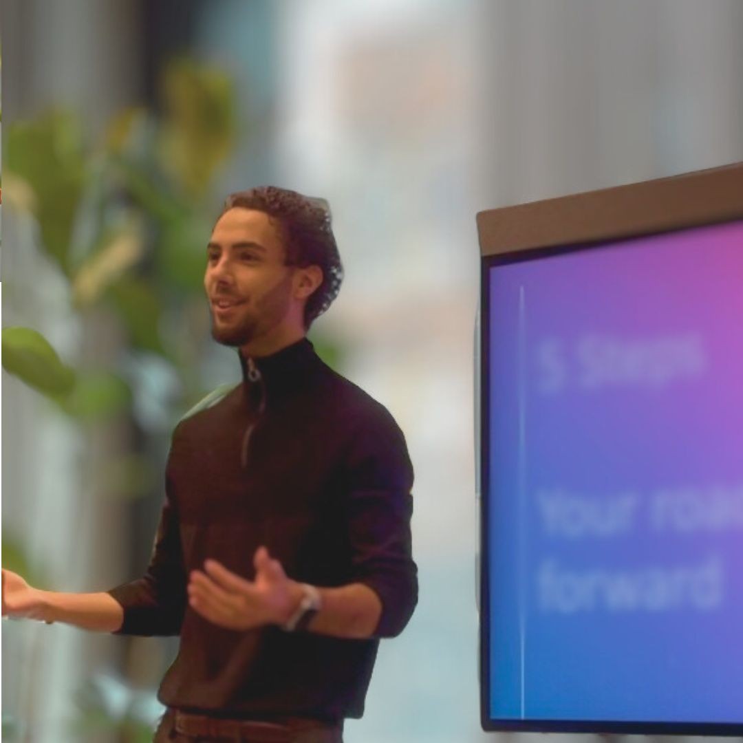 Person wearing a black sweater giving a presentation near a screen displaying a slide with blurred text.