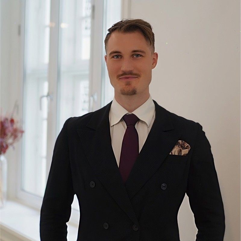 Person in a black suit and purple tie standing in front of a window with soft lighting.