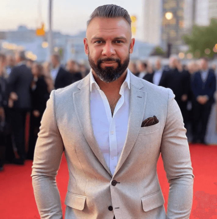 Man in beige suit standing on red carpet at event with blurred crowd in the background.