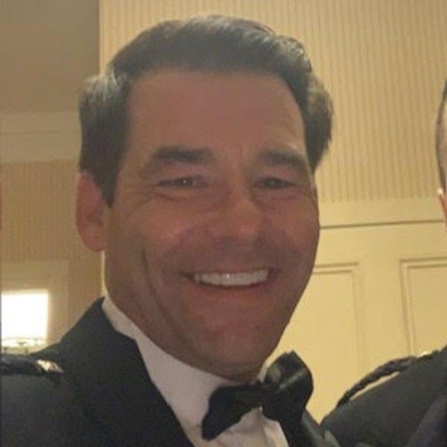 Smiling man in a tuxedo poses indoors against a light-colored wall.